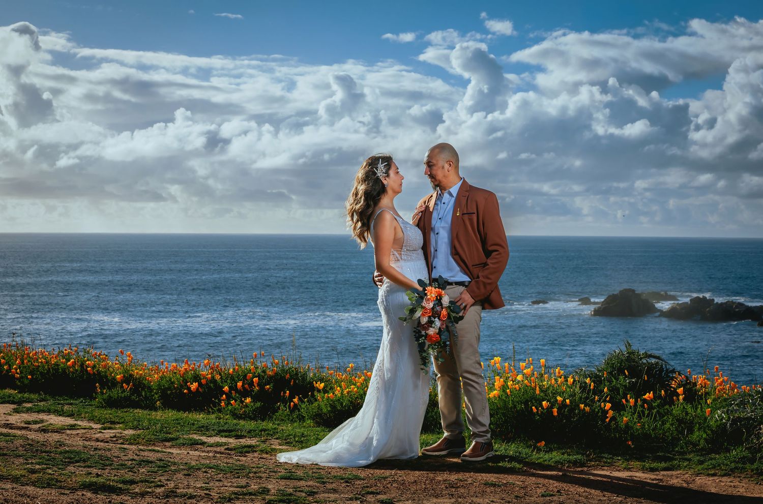 Bride in a white gown holding a bouquet and groom in a brown blazer embrace on a coastal bluff with orange wildflowers overlooking the ocean