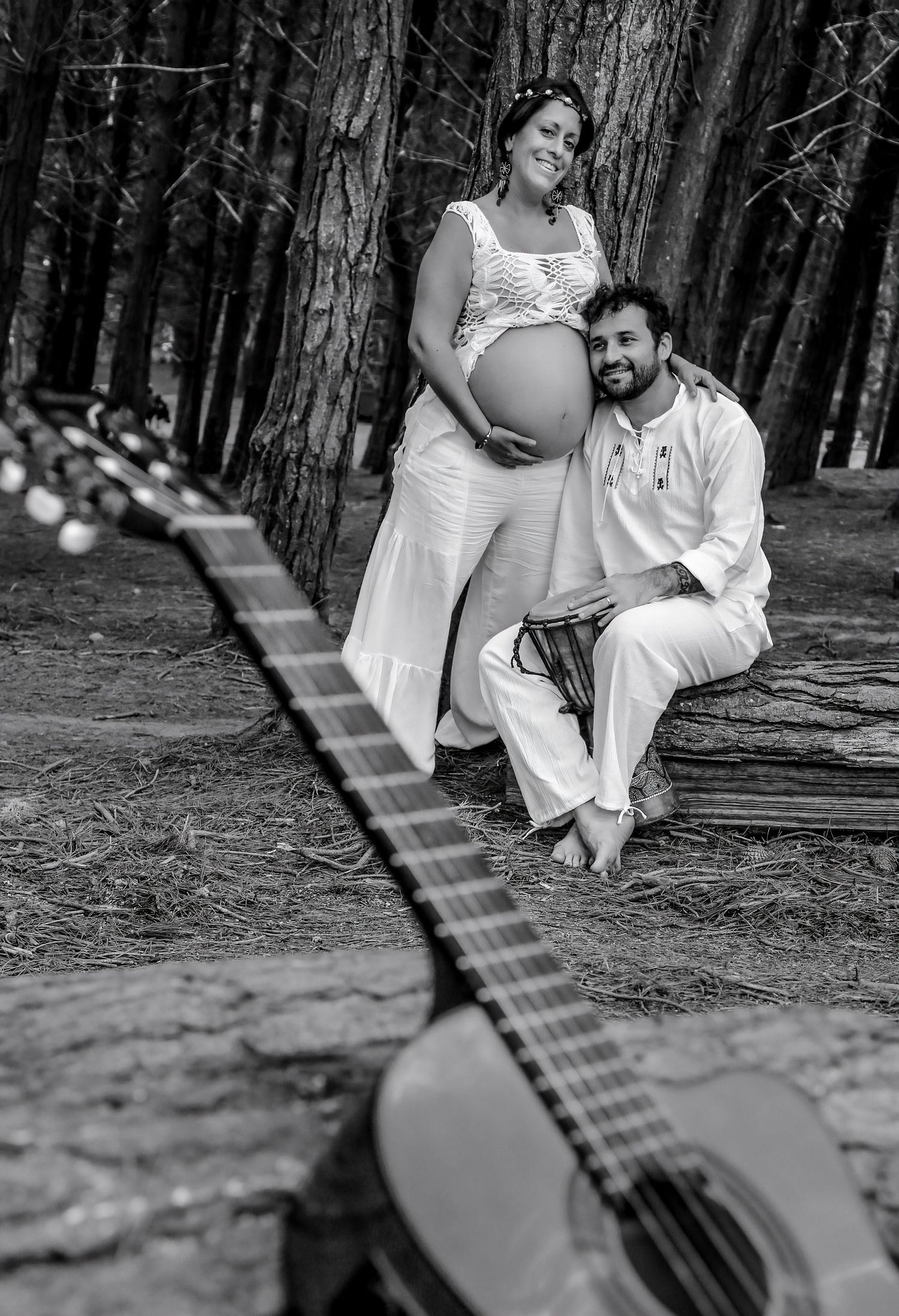 Black and white maternity photo of a couple in white outfits among trees, with a guitar in the foreground.