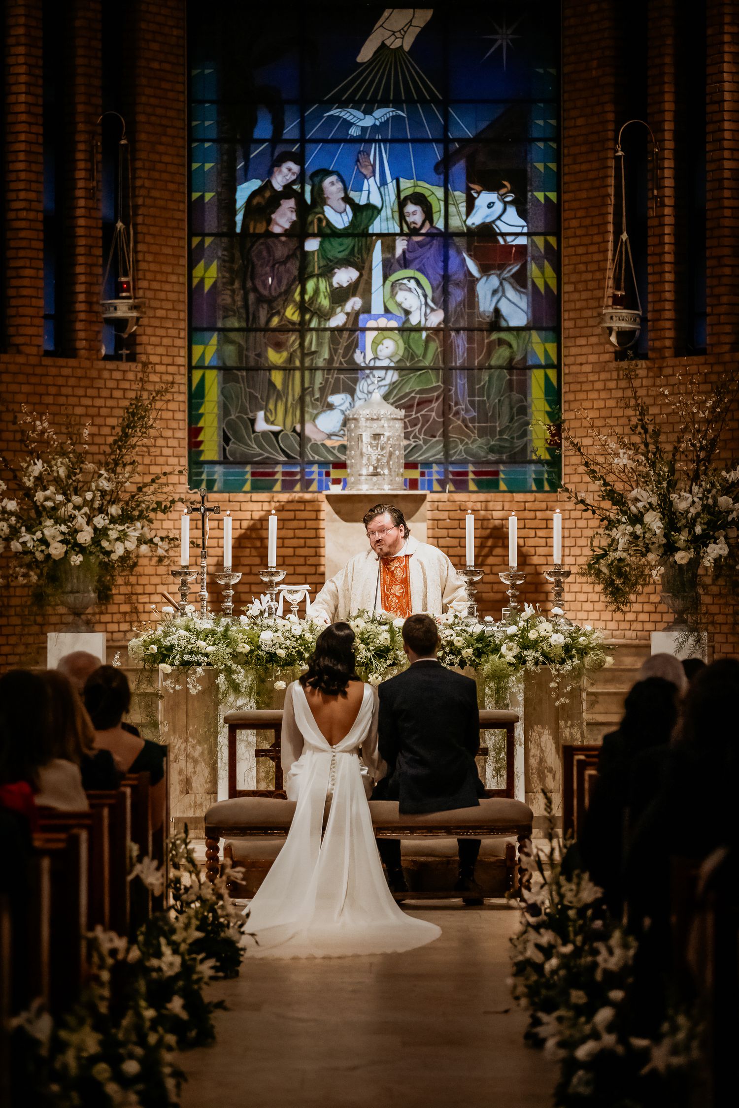 Bride and groom kneeling at church altar during wedding ceremony with stained glass window and white floral arrangements.