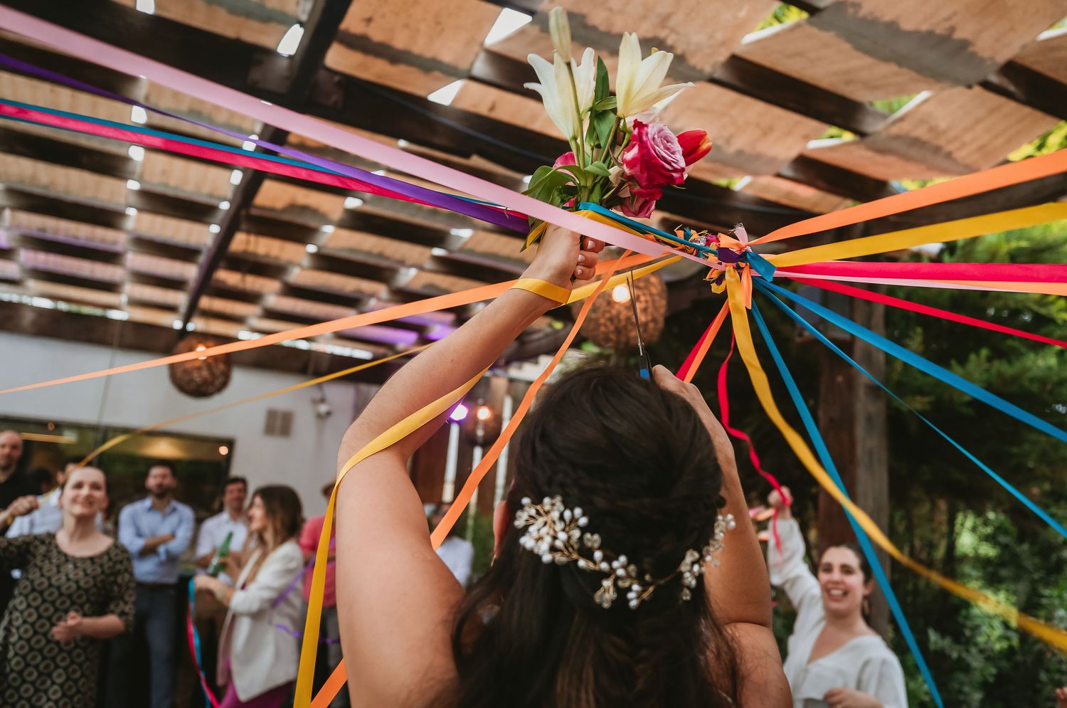 Bride tossing colorful ribbon bouquet at vibrant wedding celebration under decorated pergola.