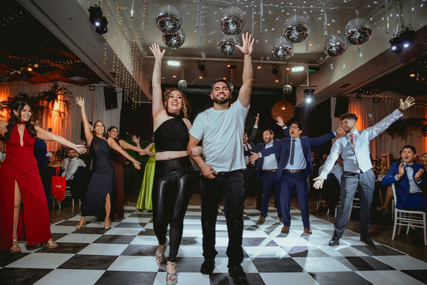 Couple leads joyful dance with arms raised on checkered floor at colorful wedding reception.