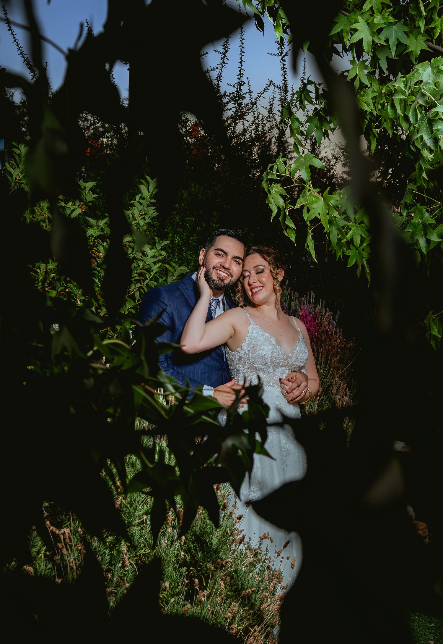 Happy wedding couple cuddles and smiles surrounded by lush green foliage in garden.