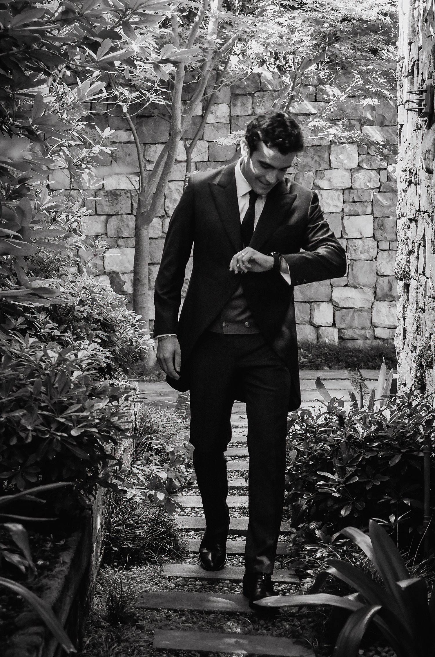 Groom in black suit checking watch while walking through lush garden pathway in black and white.