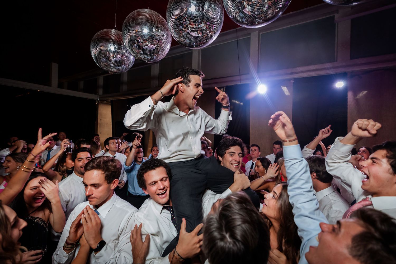 Groom crowd-surfed by cheering guests in white shirts under glowing disco balls at wedding reception.