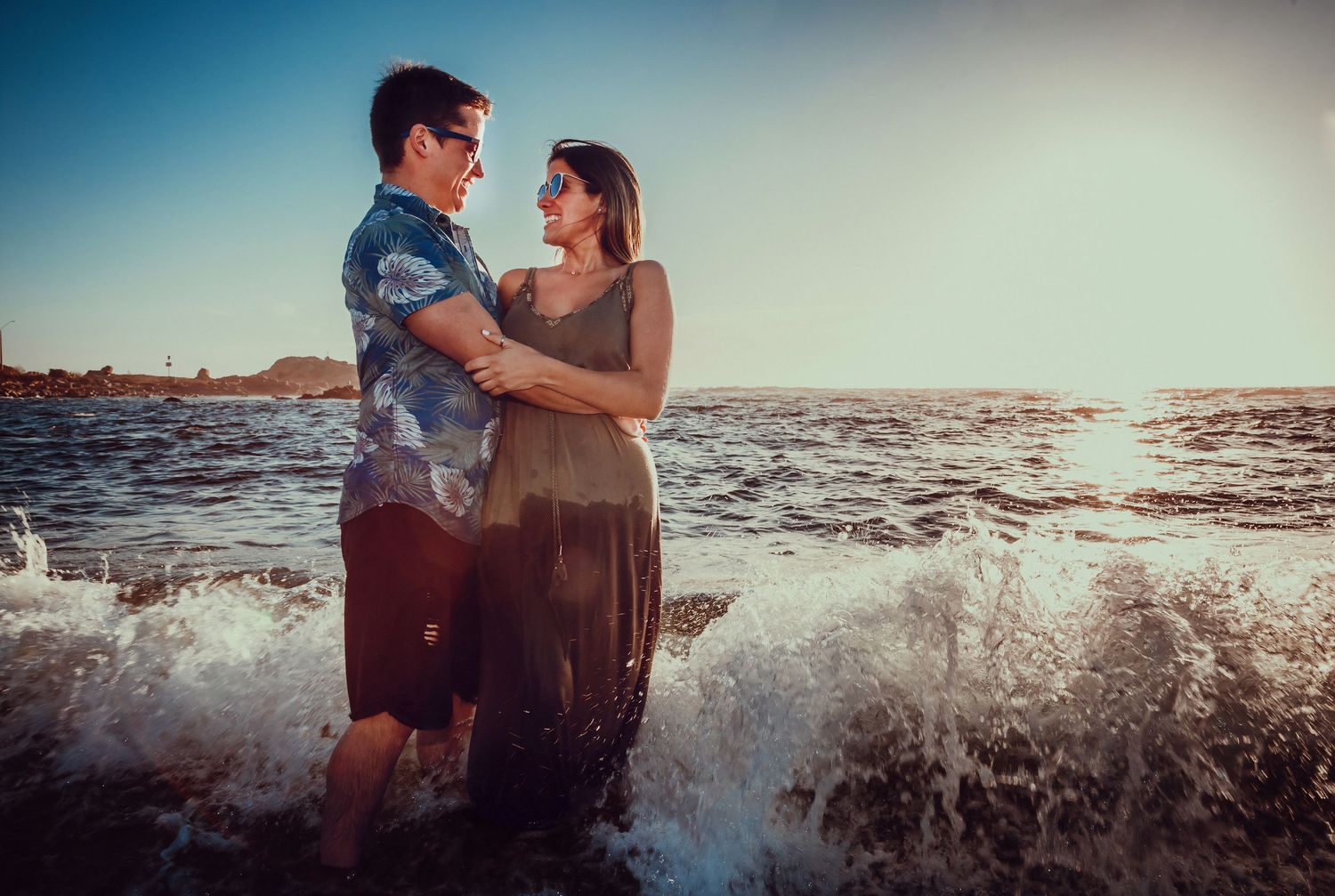 Romantic couple gazing at each other while standing in gentle ocean waves at sunset.