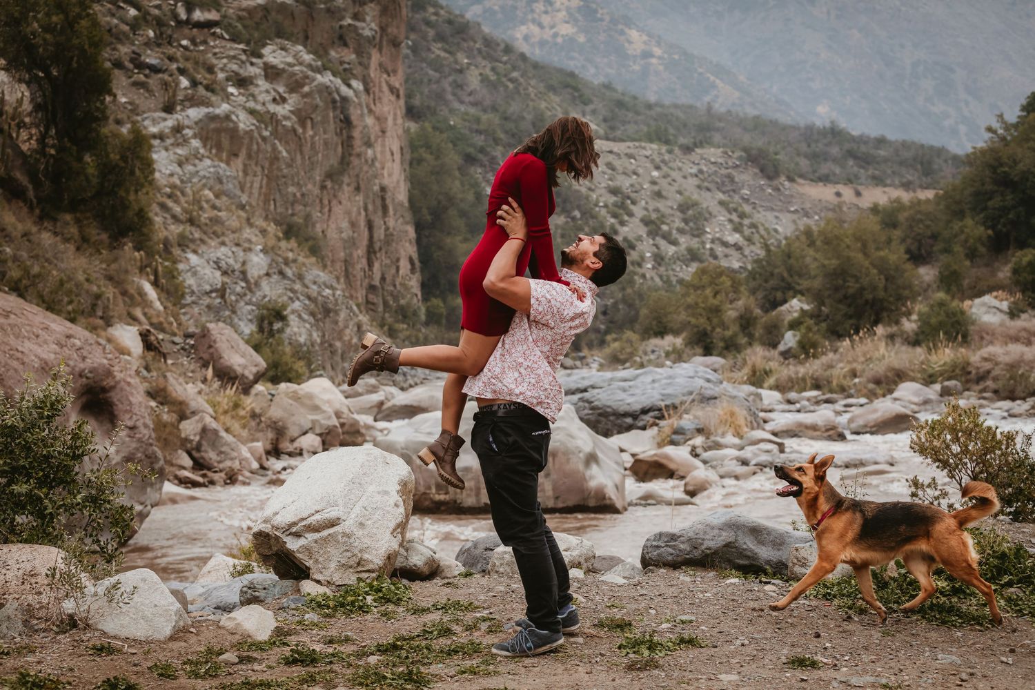 Man lifting woman in red dress joyfully outdoors with rocky mountain landscape behind them.