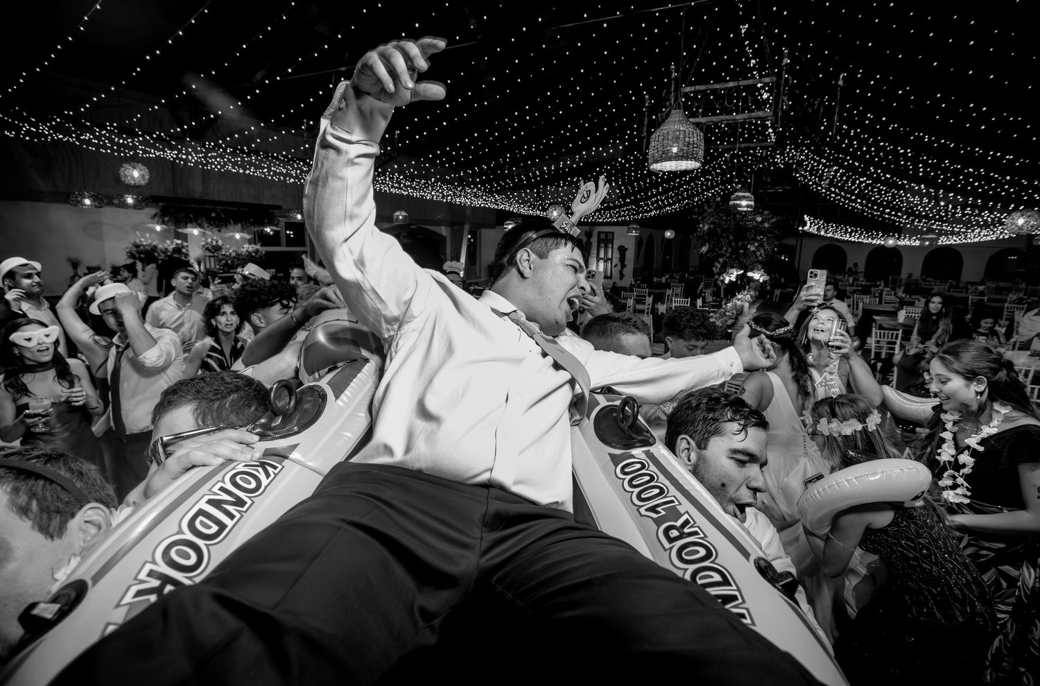 Groom crowd surfing at lively wedding reception party in black and white.