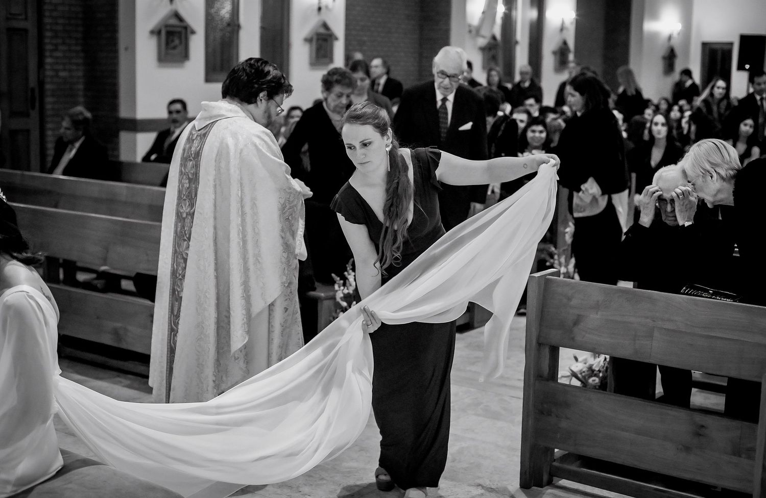 Black and white photo of a bride holding her veil during a church wedding ceremony with a priest officiating.