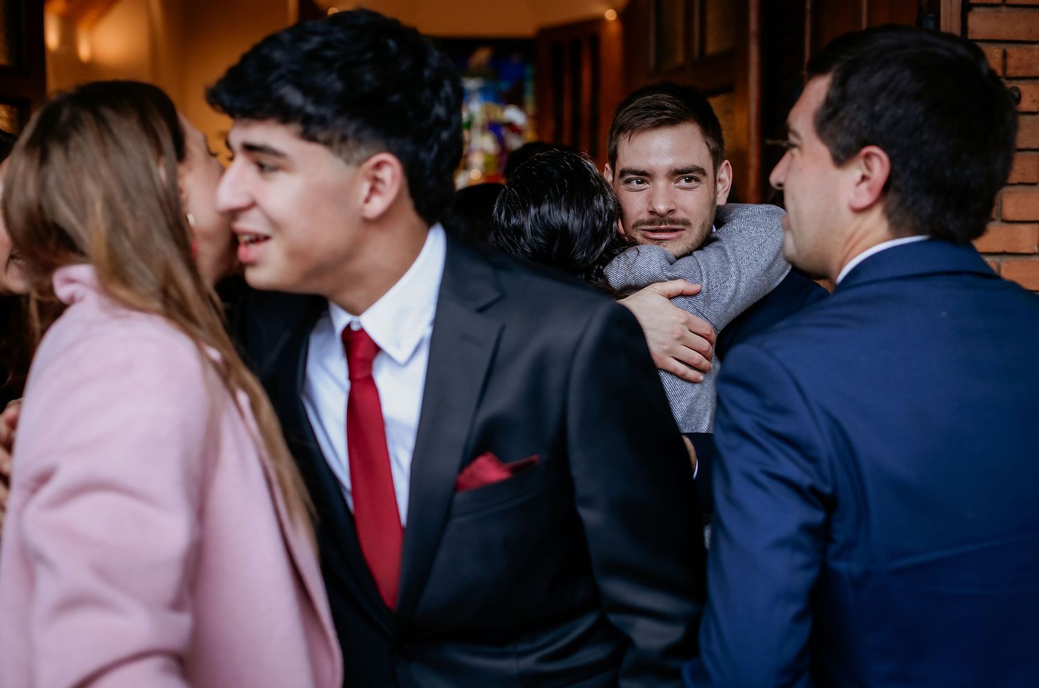Group of young men in suits socializing at a formal event, one embracing another in a warm greeting.
