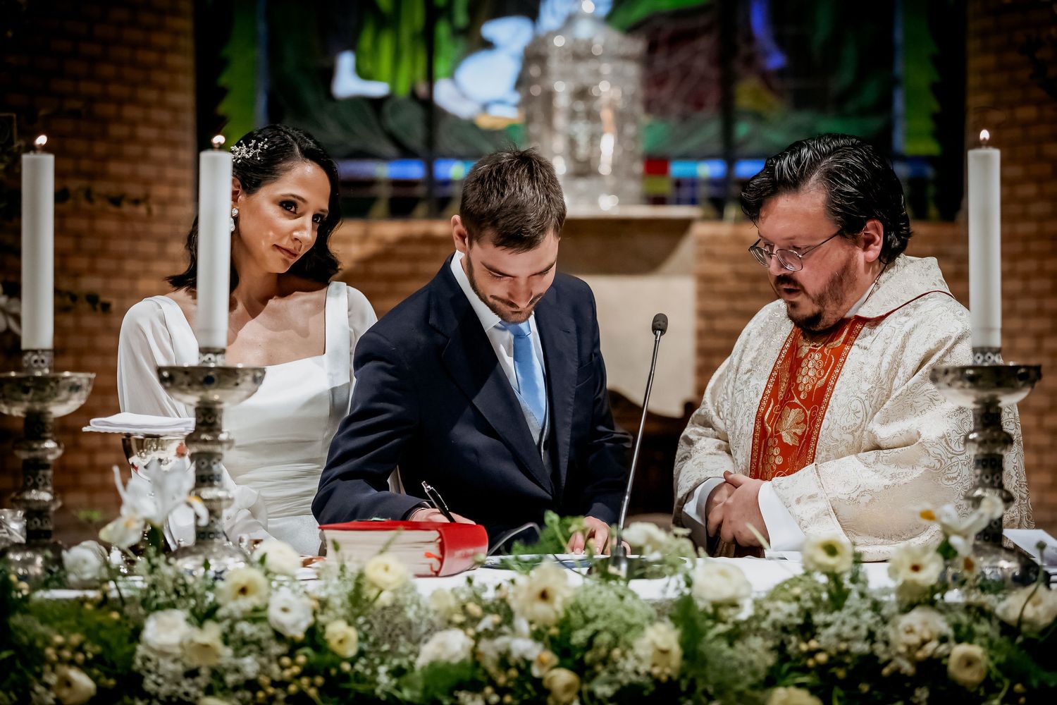 Bride and groom signing wedding documents at church altar with officiant, surrounded by white floral arrangements and candles.