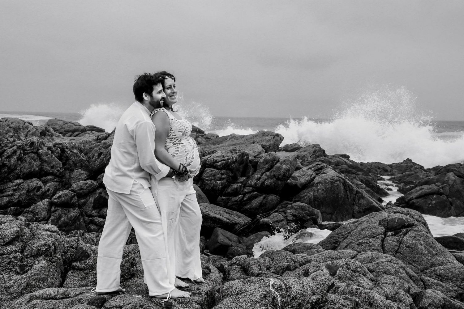 Black and white photo of a couple in wedding attire embracing on rocky shore with dramatic ocean waves crashing behind them.