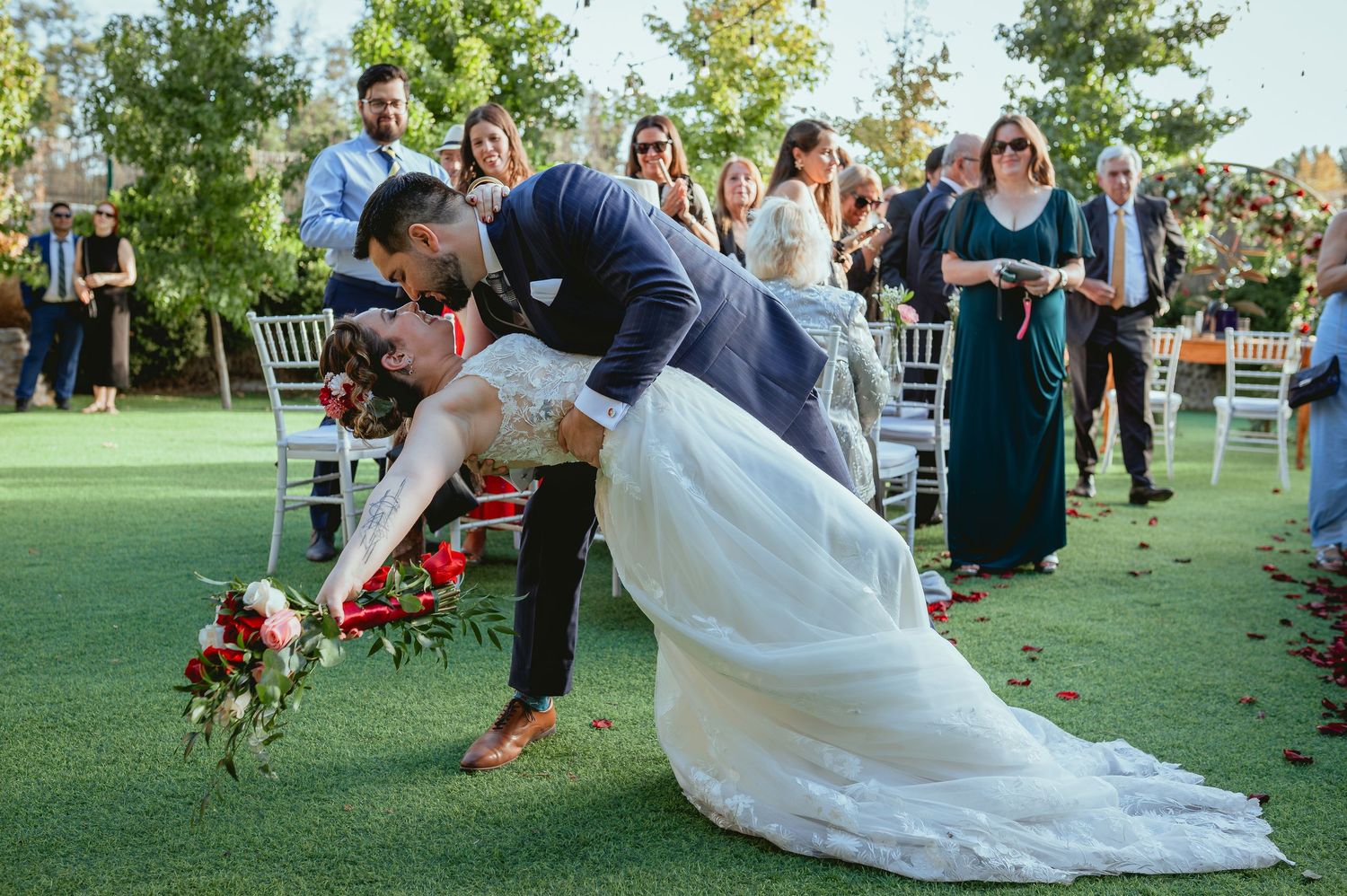Groom playfully dips bride on wedding aisle as guests cheer and celebrate at outdoor garden ceremony.