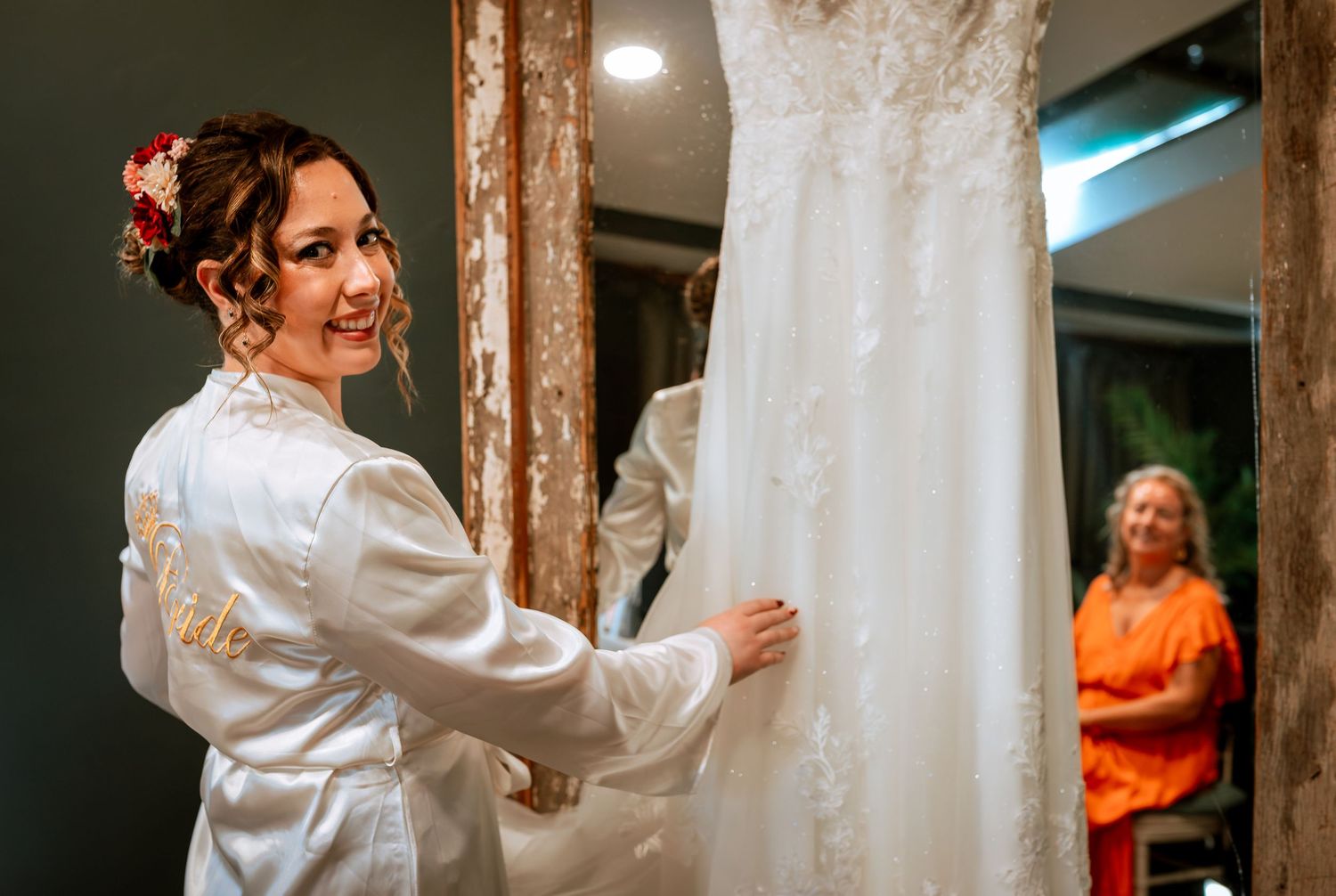 Smiling bride in embroidered robe touching lace wedding dress while woman in orange watches.