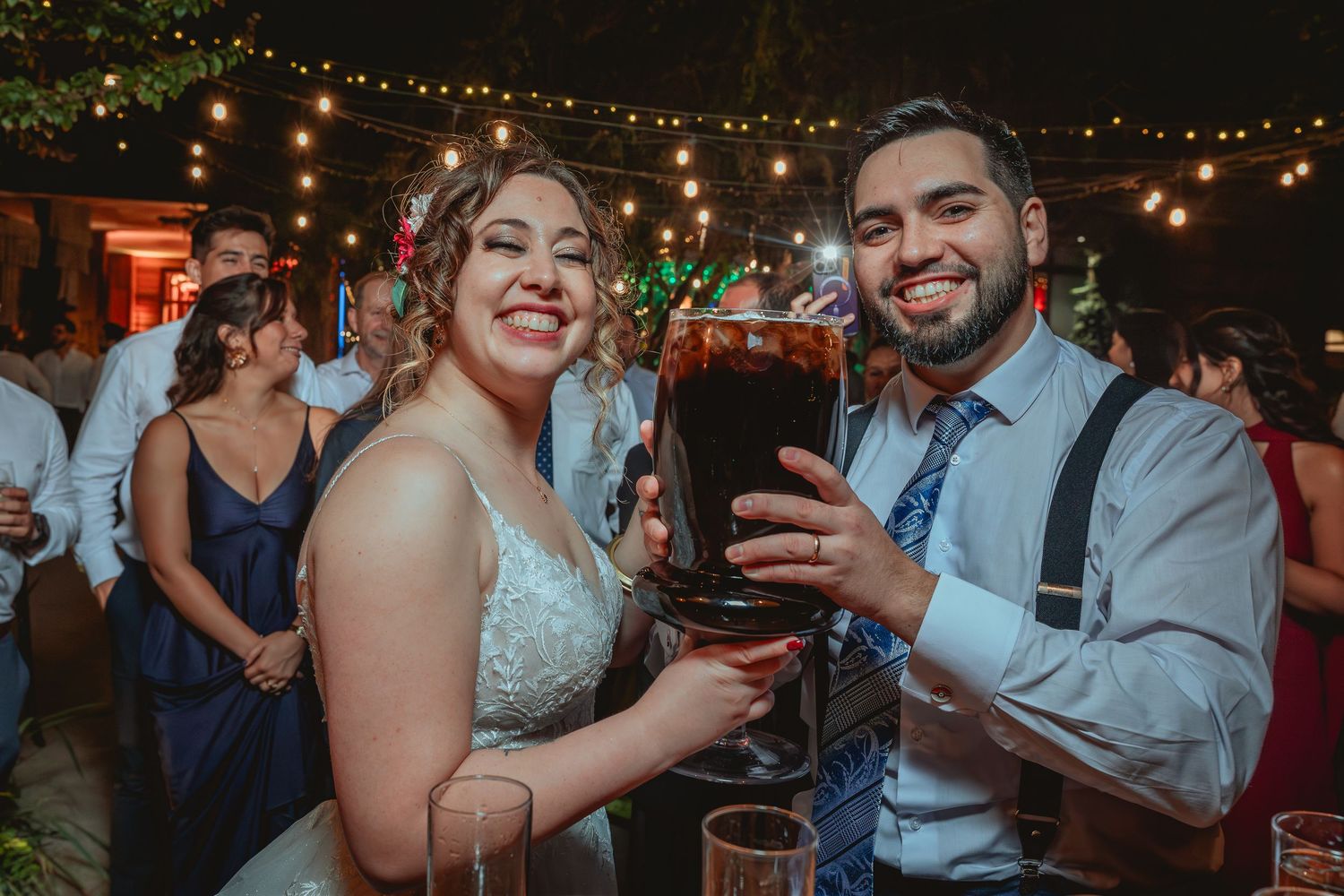Smiling bride and groom toast with oversized drink glass at outdoor wedding reception.