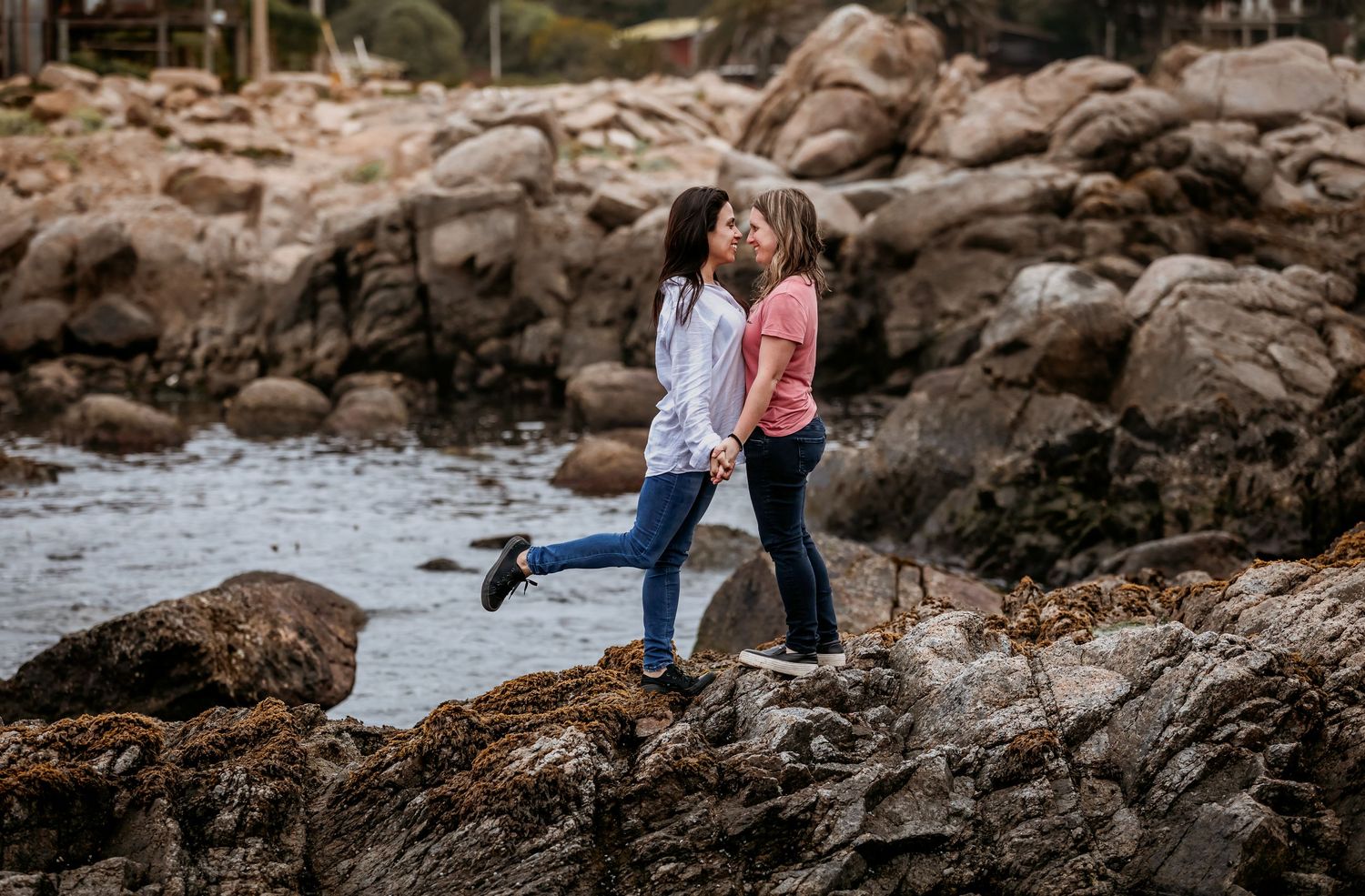Two women share a kiss while standing on coastal rocks by the ocean.