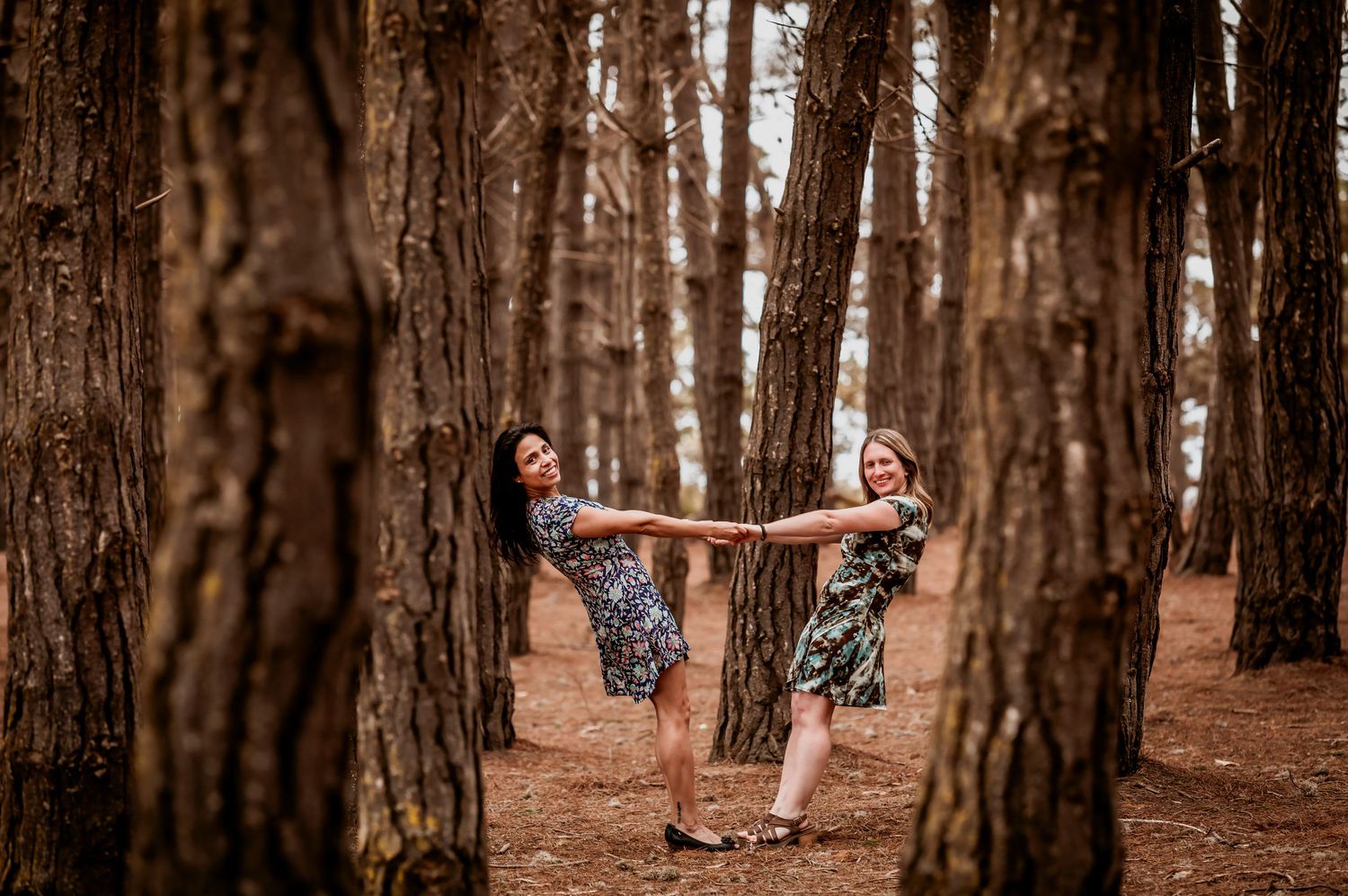 Two women playfully hold hands and lean back between tall pine trees in a forest.