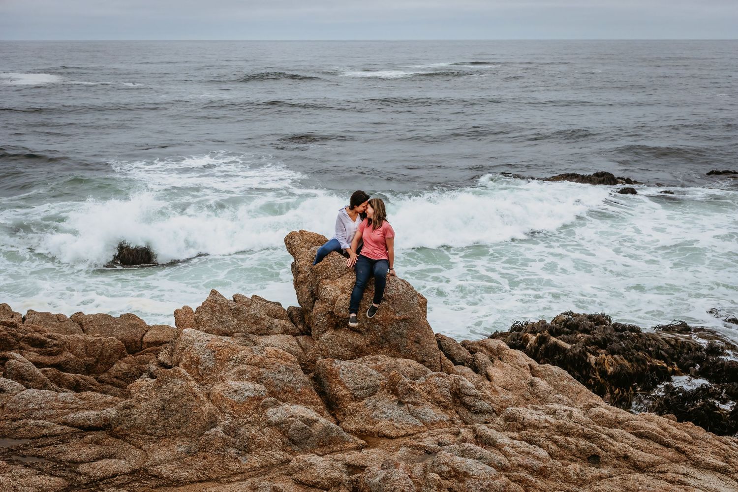Two women cuddle closely while seated on coastal rocks with crashing waves behind.