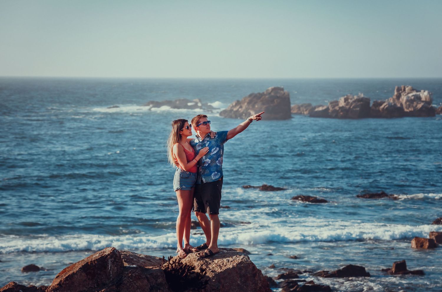 Couple standing on coastal rocks pointing at ocean view together.