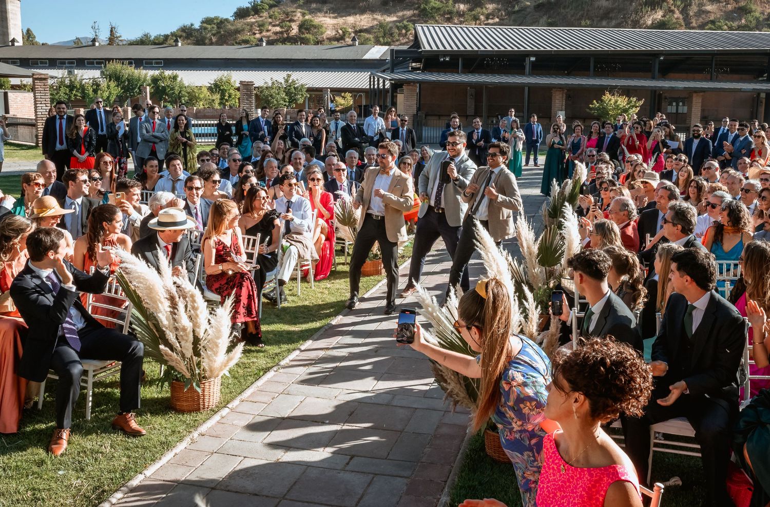 Large outdoor wedding ceremony with guests seated along an aisle decorated with pampas grass arrangements.