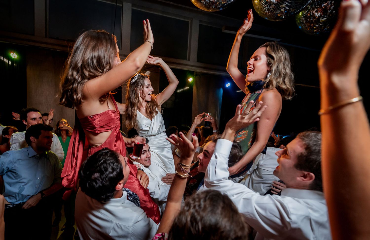 Two women in formal attire lifted above crowd of dancing guests at festive wedding reception party.