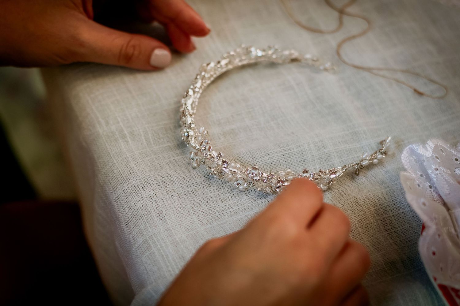 Close-up of hands carefully arranging a delicate crystal and pearl bridal tiara on white linen fabric.