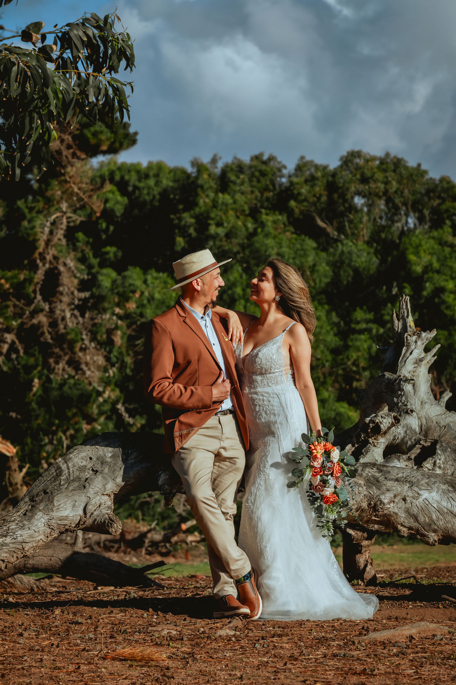 Bride in a lace wedding gown holding an orange bouquet embraces a groom in a rust blazer and straw hat near driftwood under a cloudy sky.