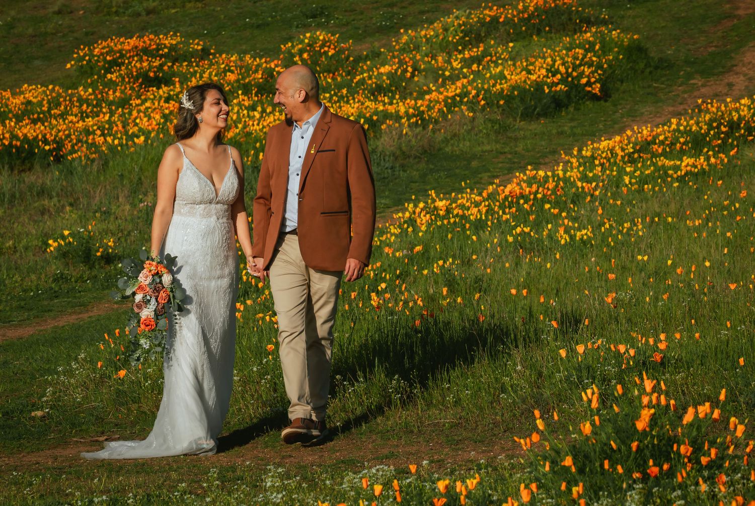 Bride in a flowing gown holding a bouquet walks hand in hand with a groom in a rust blazer through a hillside of orange wildflowers.