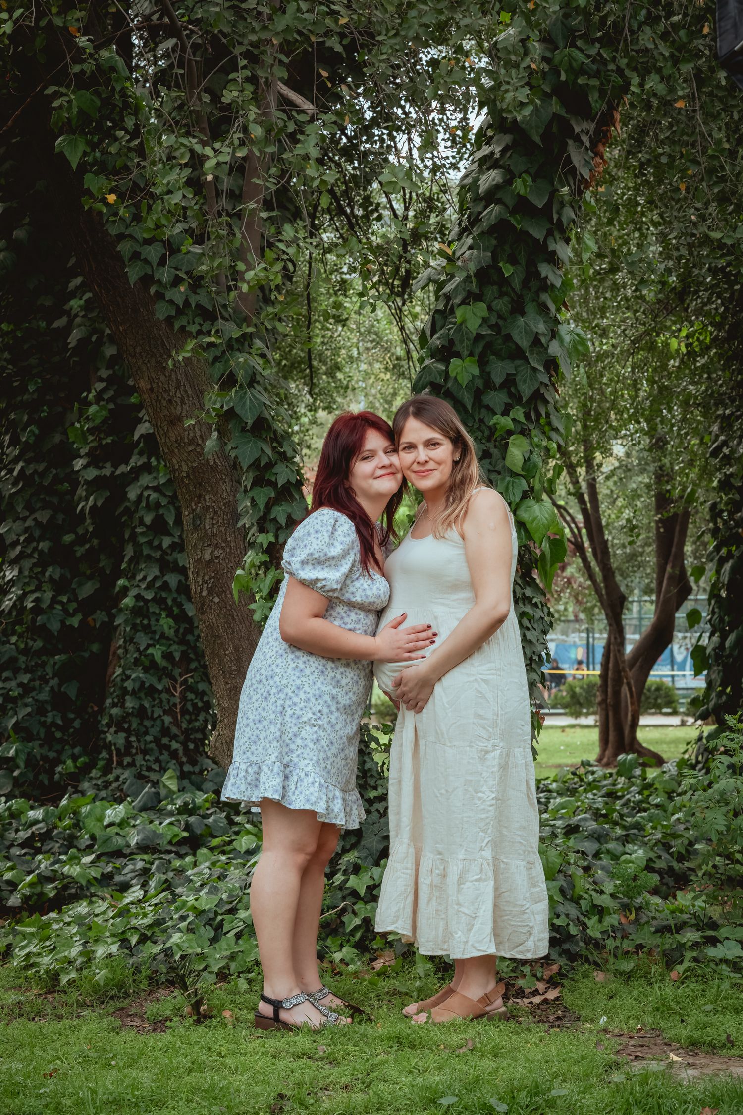 Two women in summer dresses embrace outdoors, one pregnant in white, surrounded by green ivy trees.
