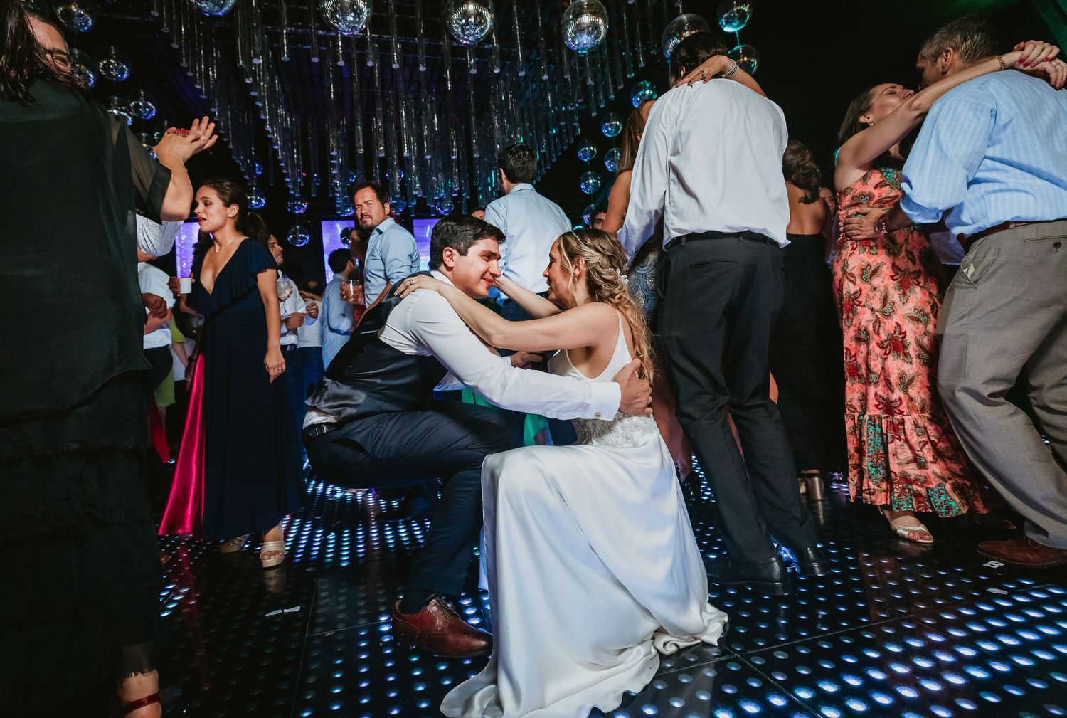 Bride and groom share playful moment on illuminated dance floor surrounded by celebrating wedding guests.