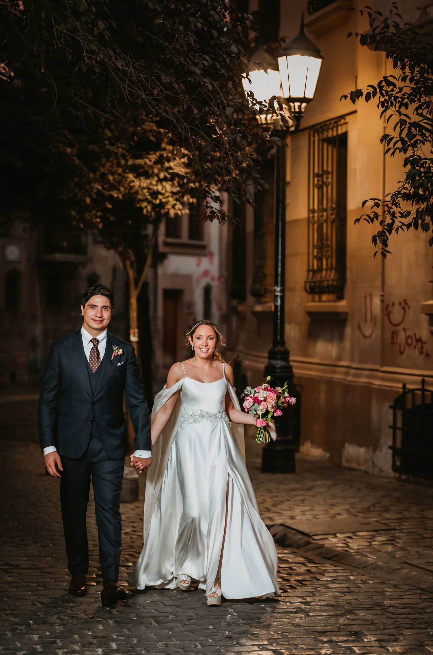 Bride and groom walking hand in hand on cobblestone street at night under warm streetlamp.