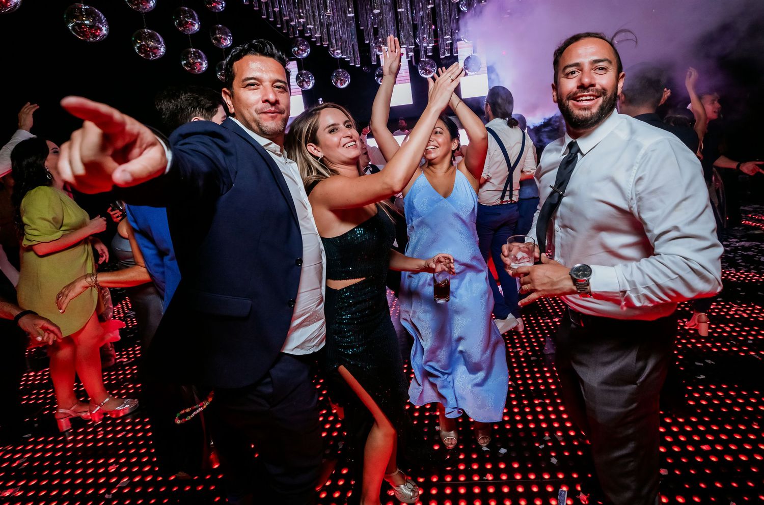 Four friends smiling and dancing together at a vibrant wedding reception party.