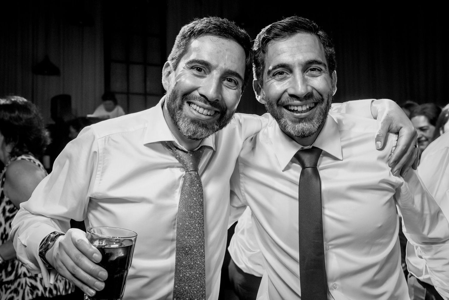 Two smiling men in dress shirts and ties pose together at a wedding reception celebration.