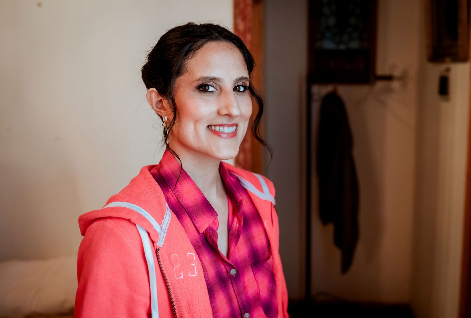 Smiling bride with updo hairstyle wearing pink hoodie and plaid shirt before wedding preparation.