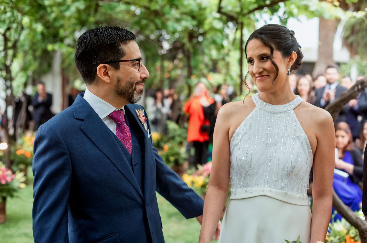 Groom in navy suit smiles lovingly at bride in beaded halter wedding gown during outdoor garden ceremony.