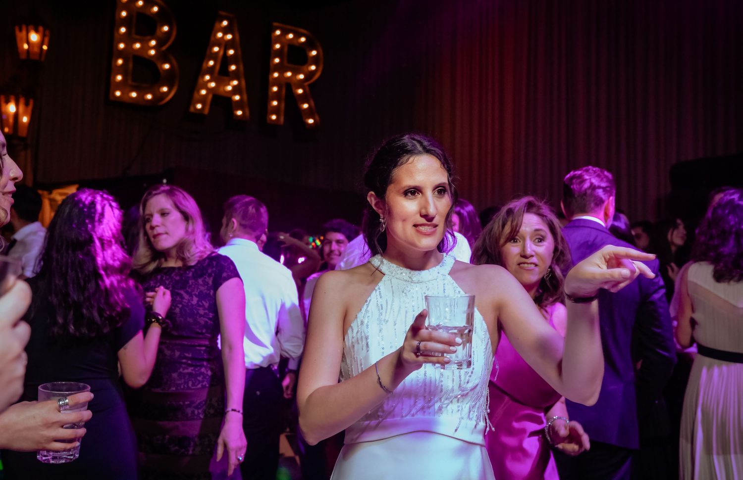 Bride in white dress holds drink while smiling at lively wedding reception under purple lights near BAR sign.