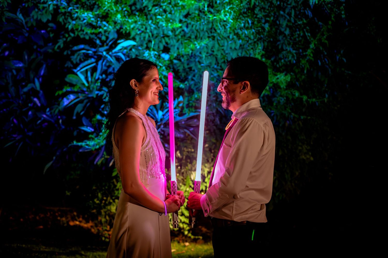 Bride and groom share a romantic moment holding glowing lightsabers in a garden at night.