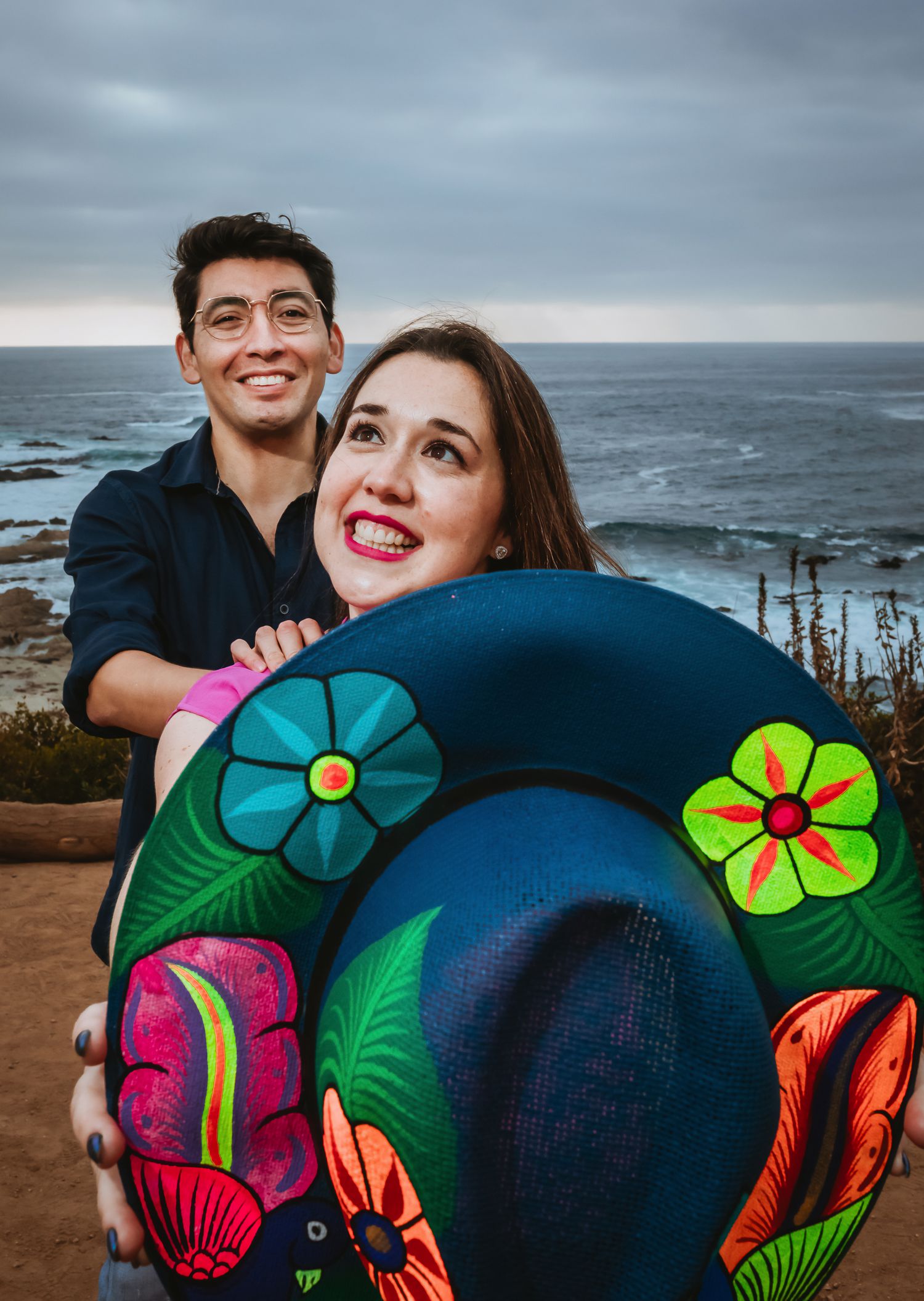 Couple smiling outdoors by the ocean, holding a colorful floral painted sombrero hat near a coastal cliff.