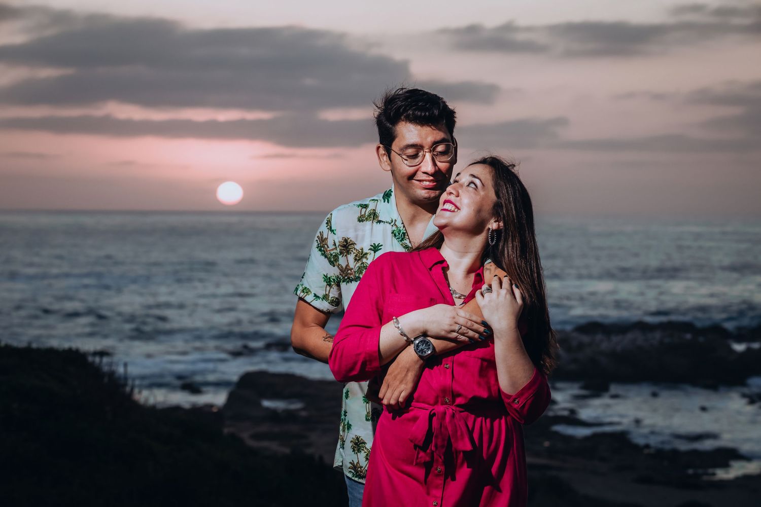 Couple embracing at sunset by the ocean, woman in red dress smiling up at man in floral shirt, romantic beach photography.