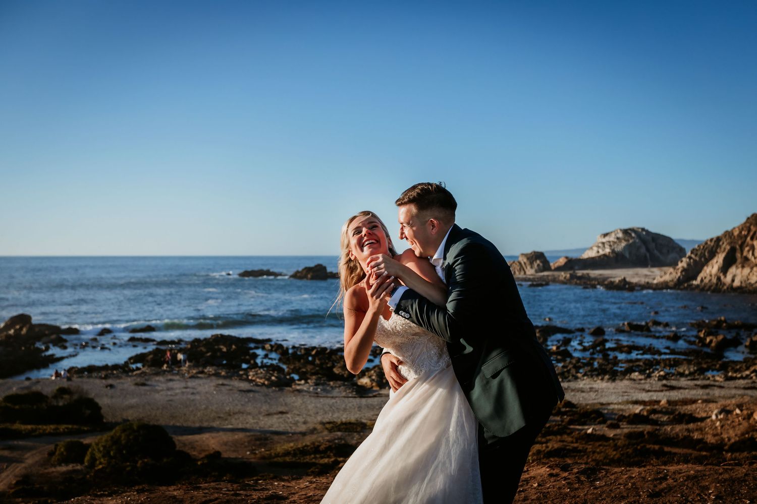Bride and groom share playful moment dancing together on rocky California coastal beach at sunset.