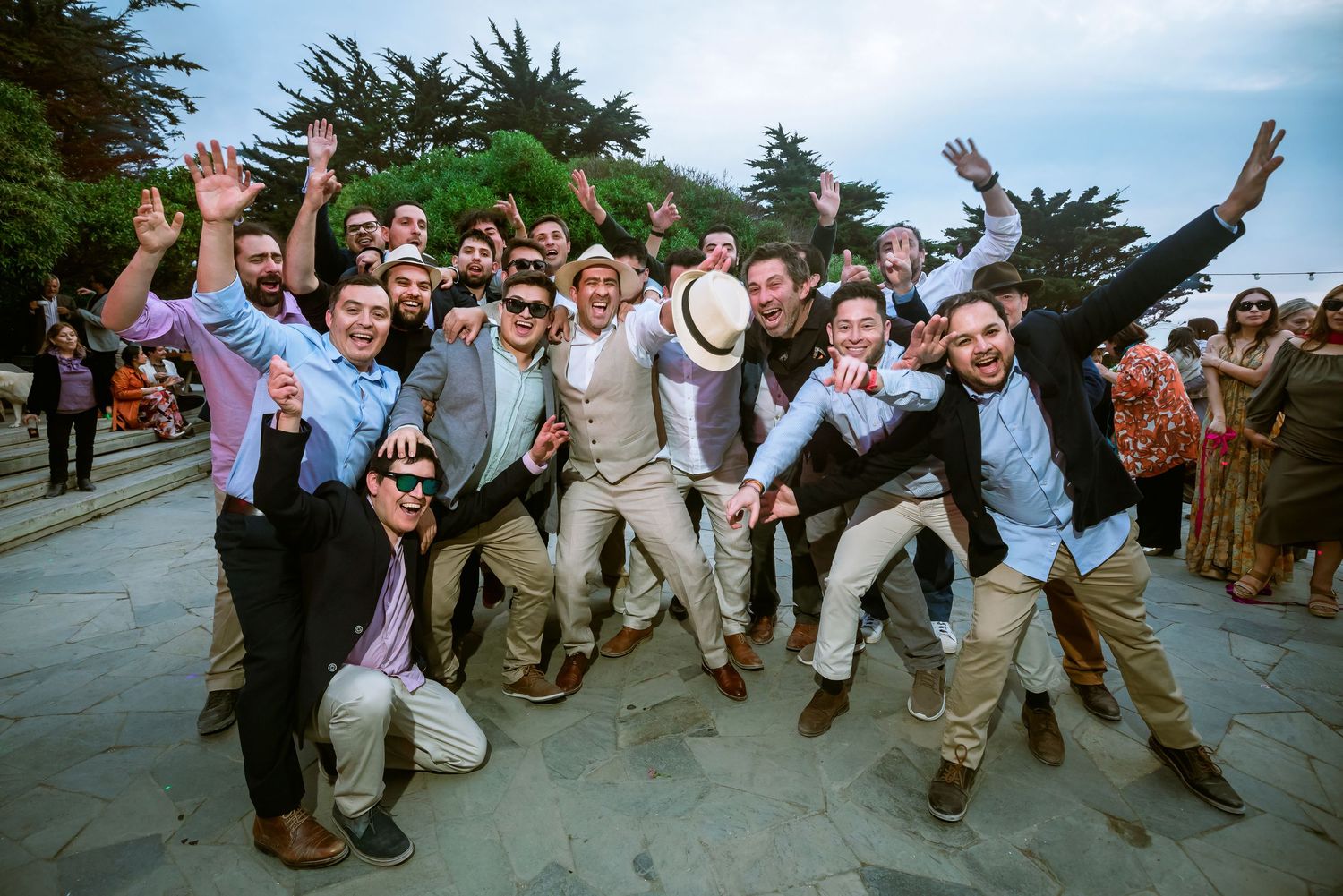 Group of men celebrating joyfully outdoors at a wedding reception, jumping and posing for a fun group photo.