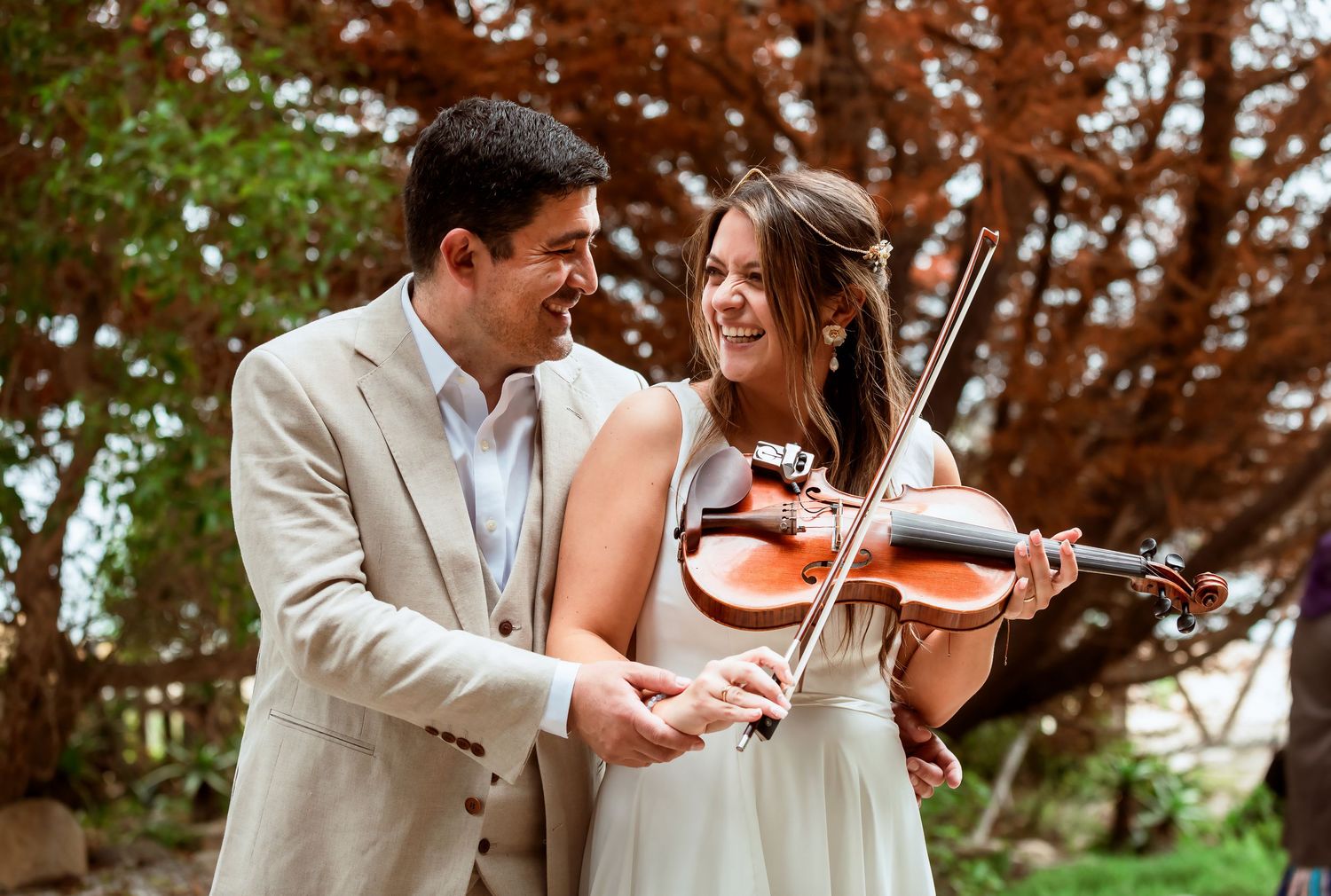Smiling groom in beige suit stands beside bride playing violin at an outdoor wedding celebration.