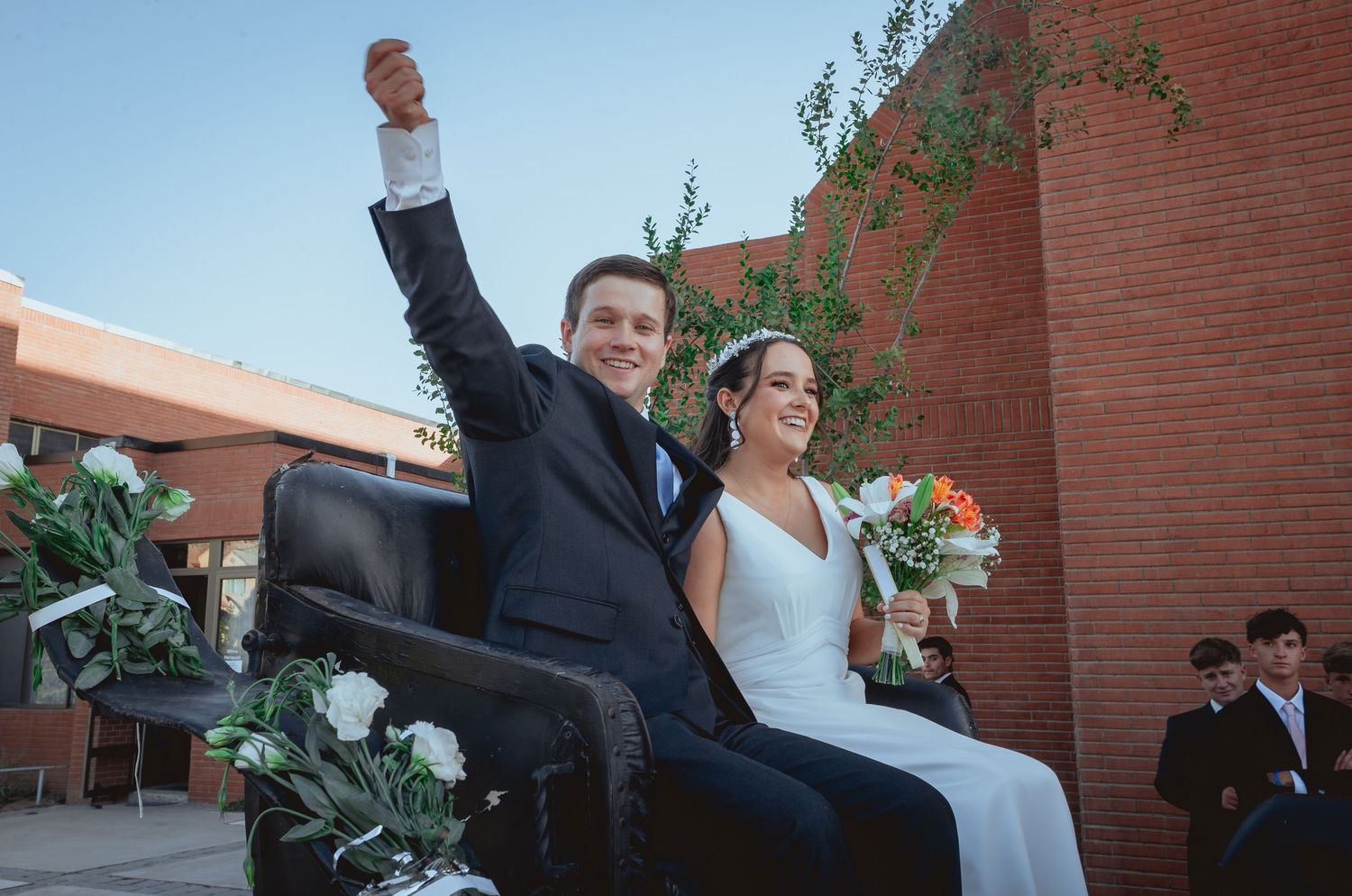 Joyful newlyweds ride in vintage car, groom raising fist in celebration after wedding.