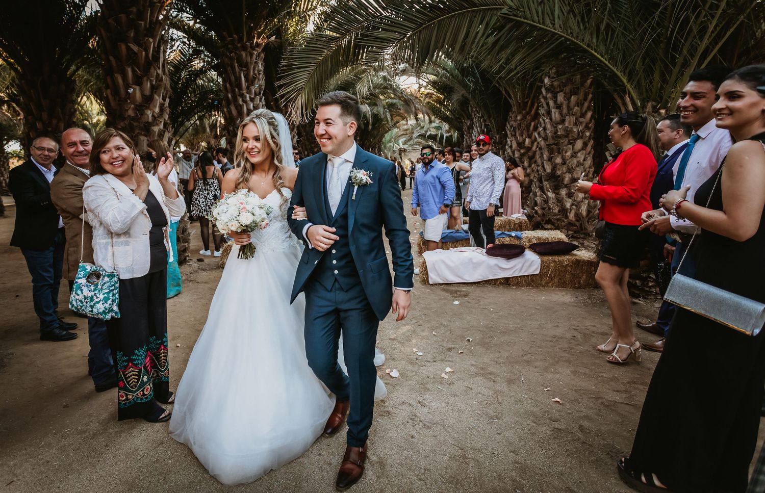 Smiling couple walks hand-in-hand past guests after outdoor wedding ceremony among tall palm trees.