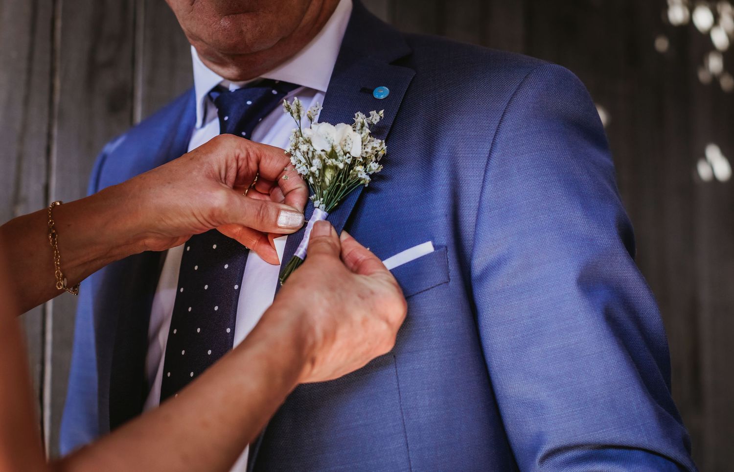 Hands pinning white floral boutonniere on groom in blue suit with polka dot tie.