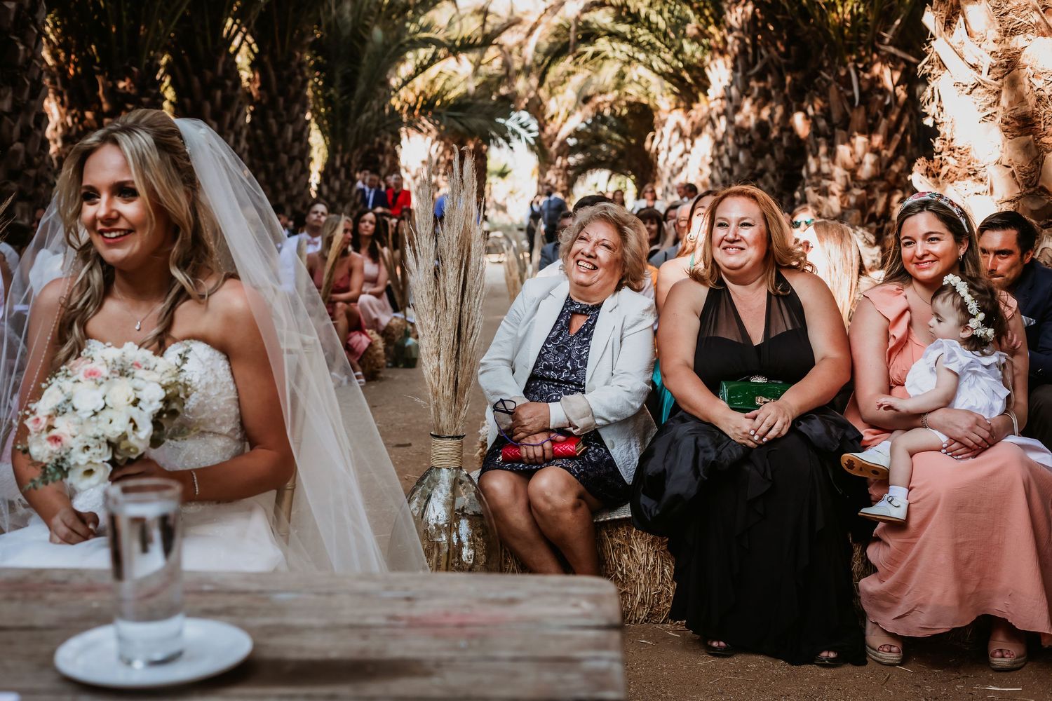 Bride in white gown and veil holds bouquet while smiling guests watch outdoor wedding ceremony.