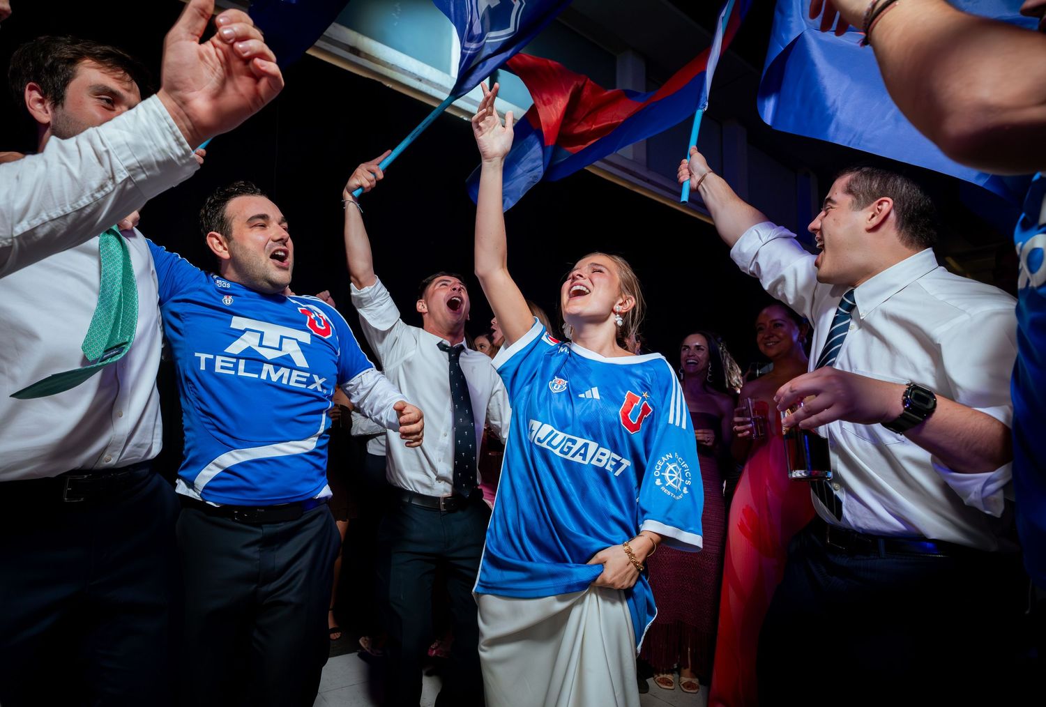 Wedding guests celebrate in blue soccer jerseys, waving flags at festive reception.