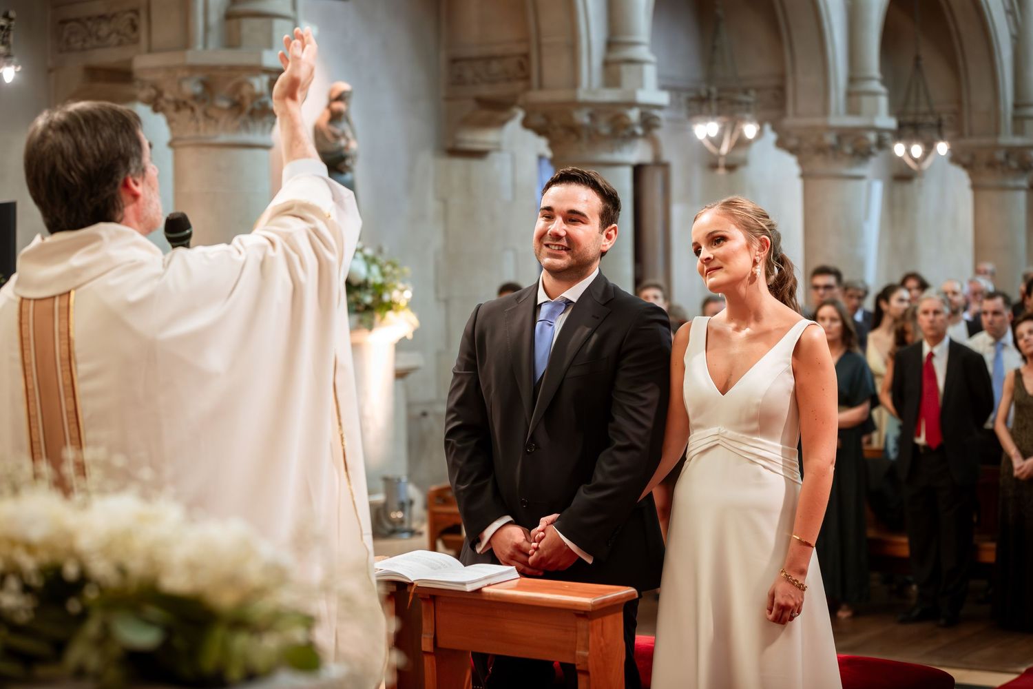 Priest blesses couple during Catholic wedding ceremony at church altar with guests watching.