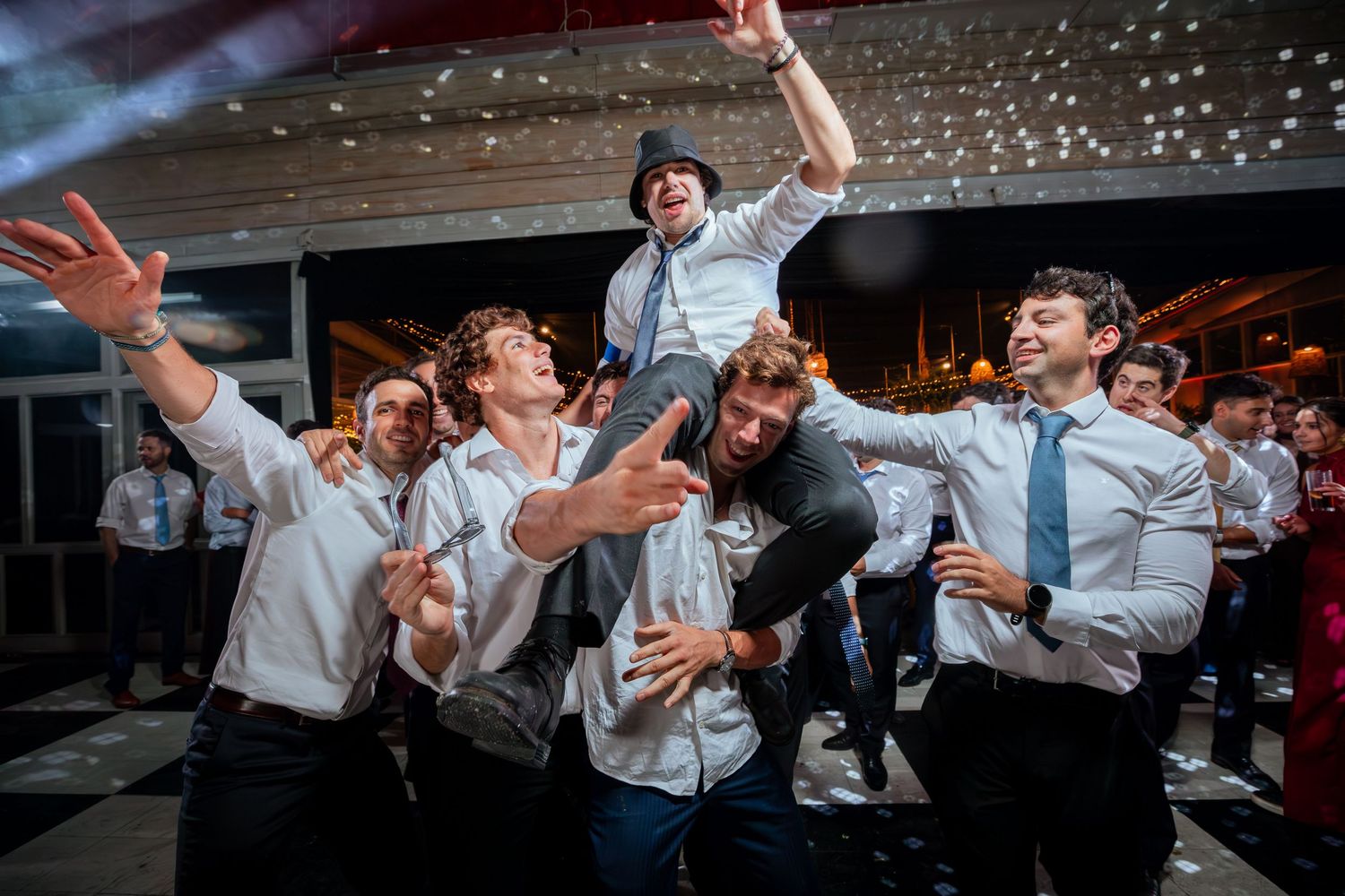 Groomsmen in white shirts lift and carry friend on shoulders at wedding.