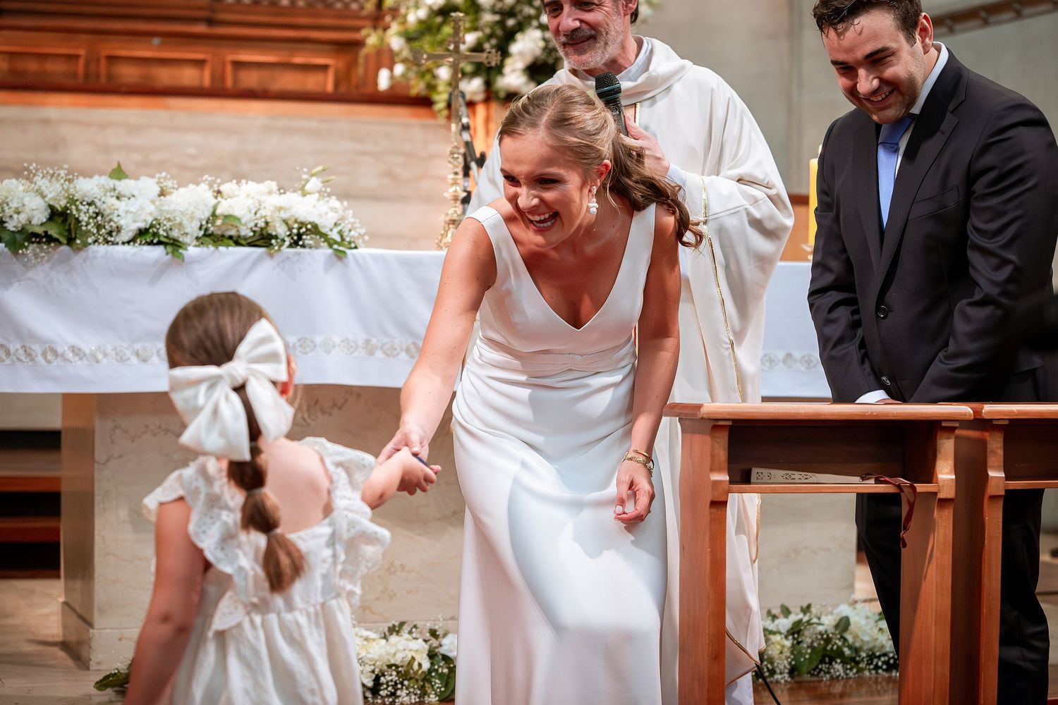 Bride in white gown bends to greet flower girl in white dress at church altar.