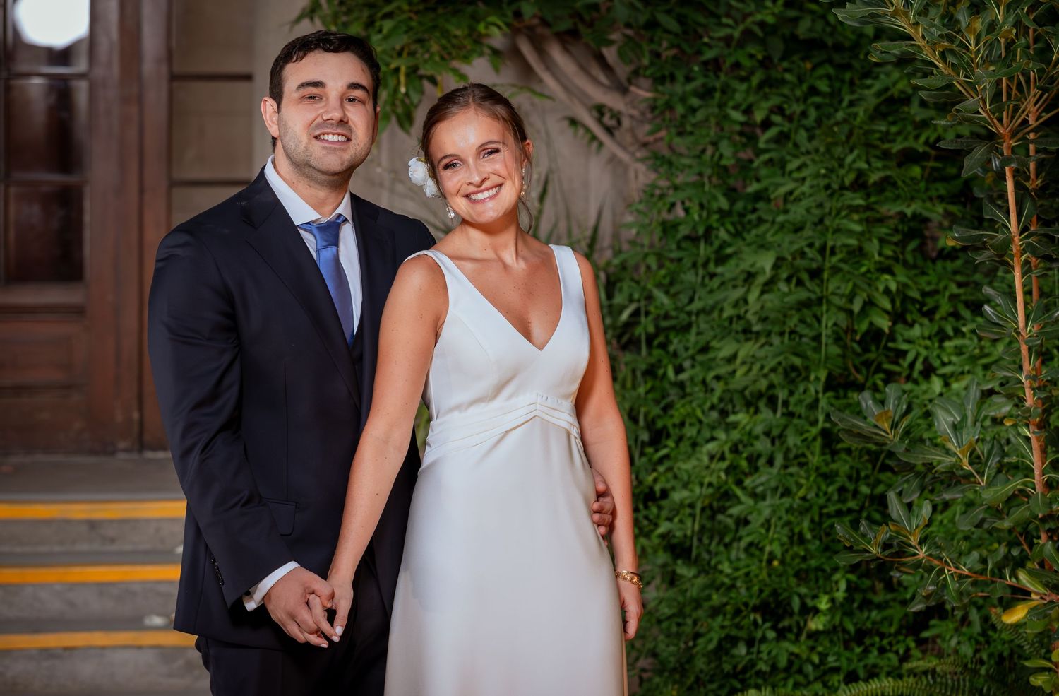 Happy bride and groom hold hands while posing in front of lush green garden wall.